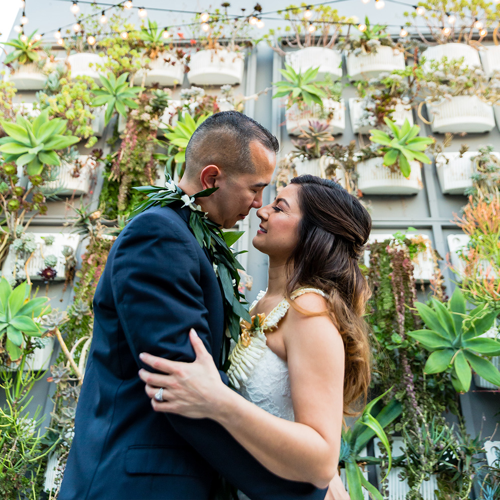 A happy couple holding each other in front of a wall of succulents on The Colony House patio