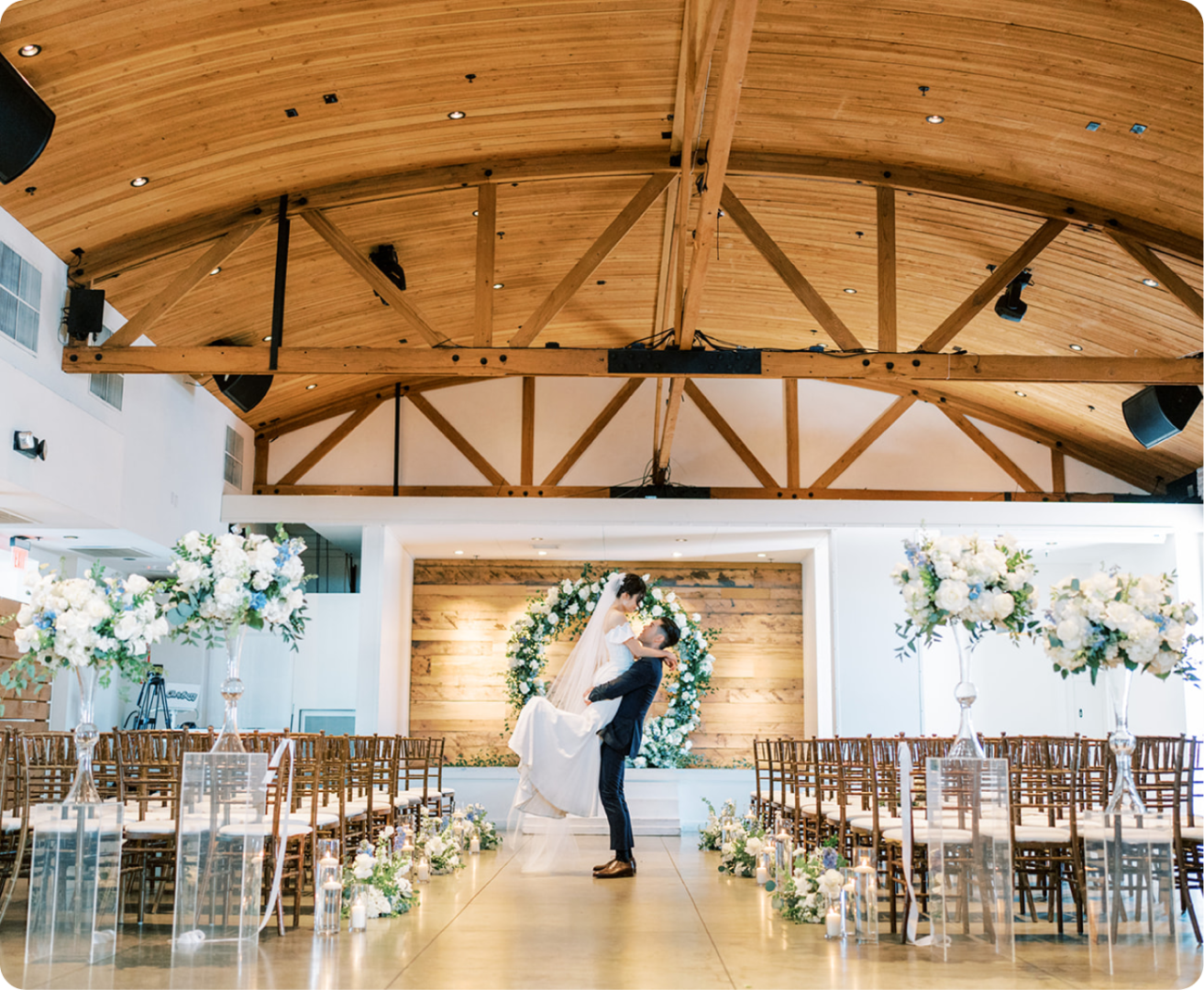 A groom lifts his bride in the center aisle of a bright wedding venue, surrounded by empty chairs and large floral arrangements, beneath a wooden vaulted ceiling.