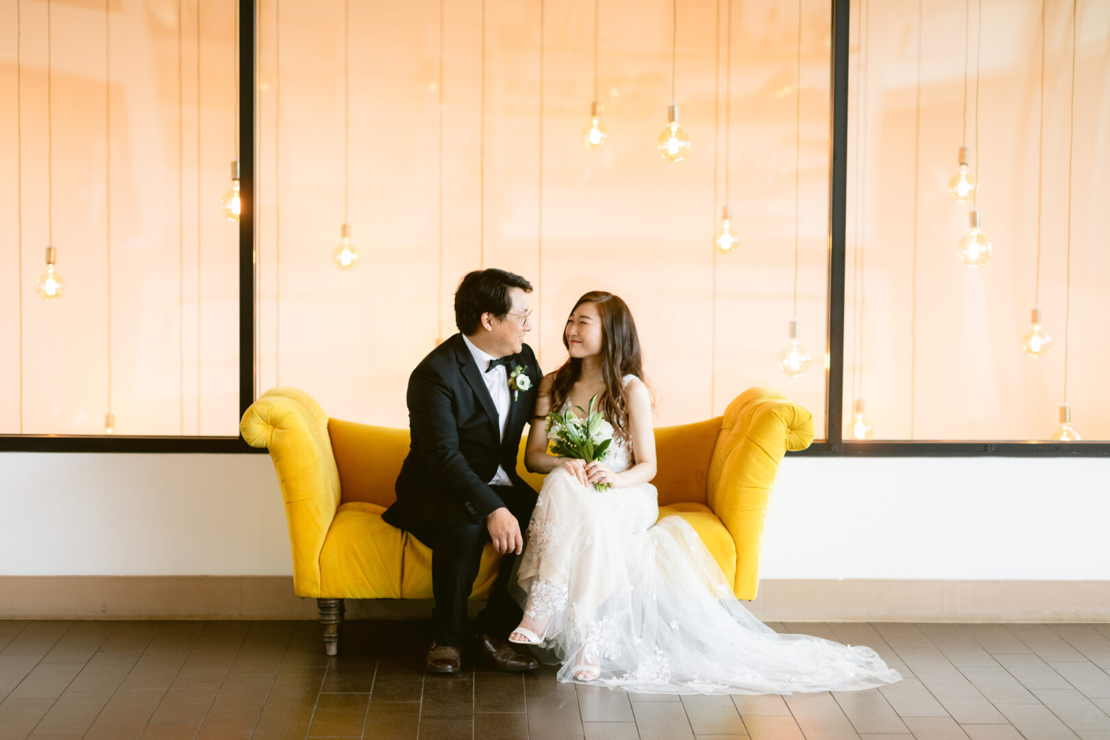 A couple in wedding attire sits on a bright yellow couch, smiling at each other. Soft lights hang in the background, creating a warm and romantic atmosphere.