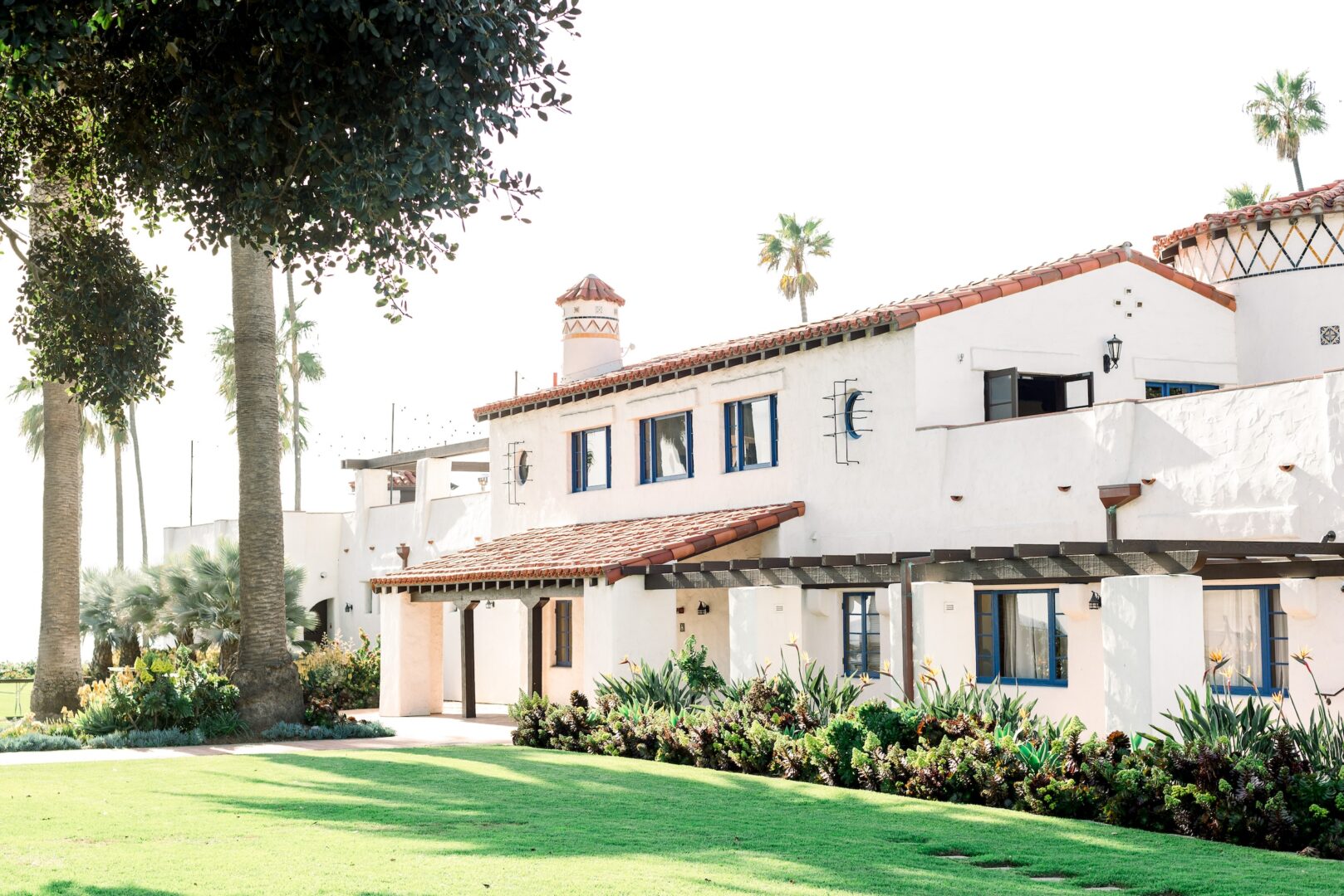 A white Spanish-style building with a red tile roof is surrounded by palm trees, green lawn, and lush landscaping on a sunny day.