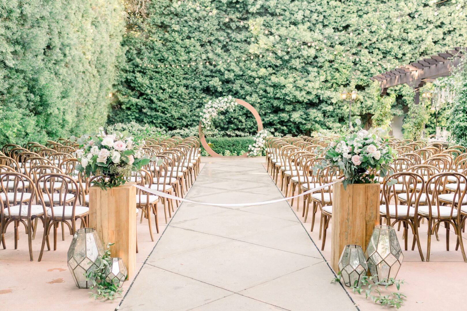 An outdoor wedding ceremony setup with rows of wooden chairs, floral arrangements, lanterns, and a circular floral arch at the end of the aisle, surrounded by lush greenery.