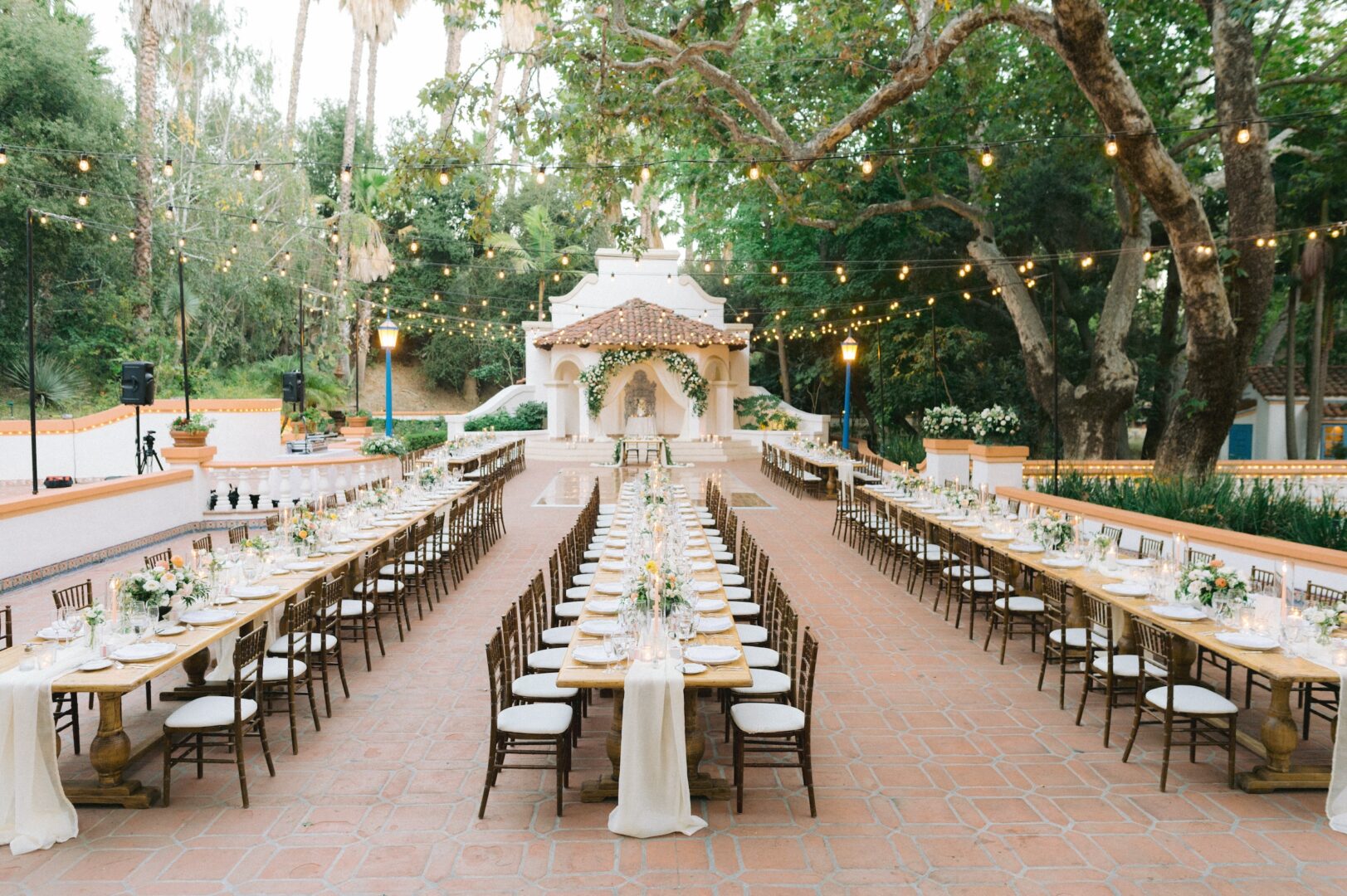 Outdoor wedding reception setup with two long dining tables, floral centerpieces, wooden chairs, string lights overhead, and a decorated altar at the front, surrounded by trees and greenery.