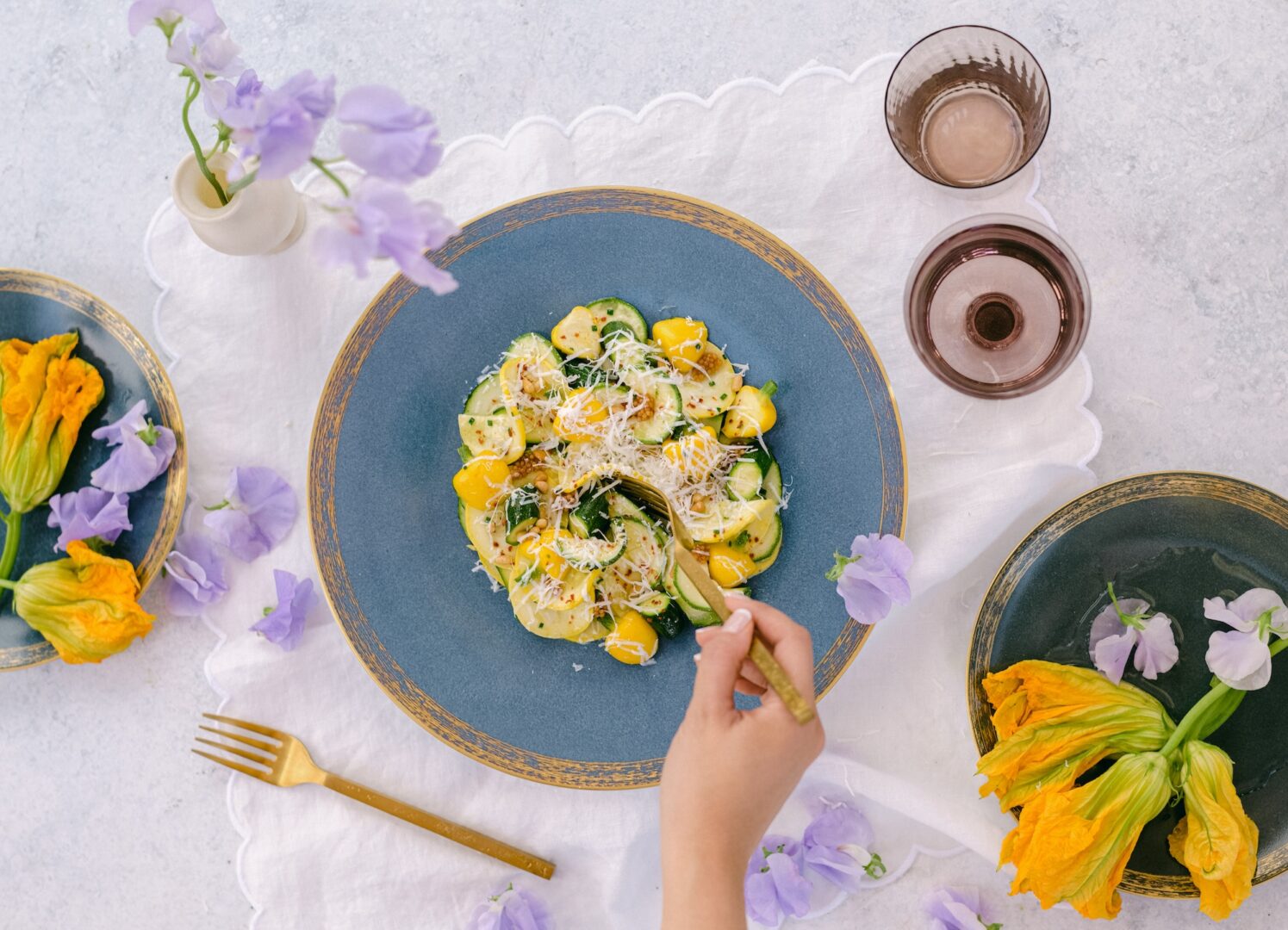 A hand holds a fork over a blue plate of pasta with yellow squash, zucchini, and grated cheese, surrounded by flowers, a gold fork, and a glass on a white tablecloth.