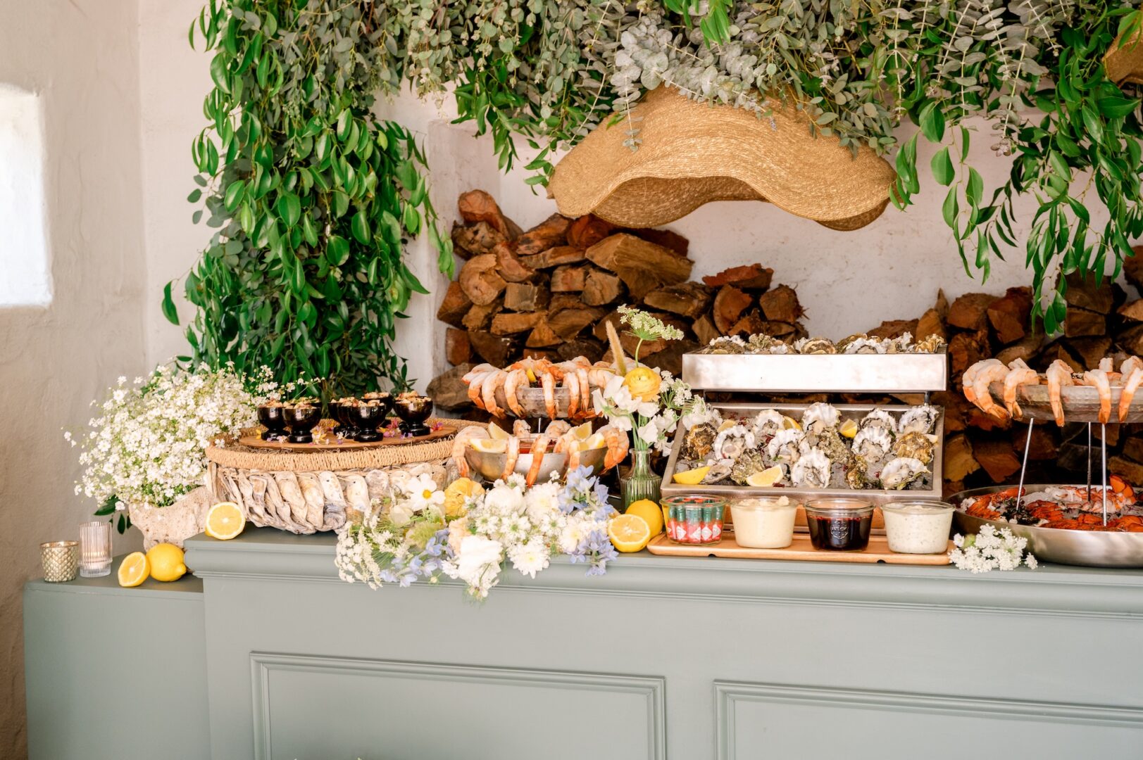 A seafood buffet with shrimp, oysters, and dipping sauces is displayed on a green counter, decorated with lemons, flowers, and greenery, with stacked firewood and hanging leaves in the background.