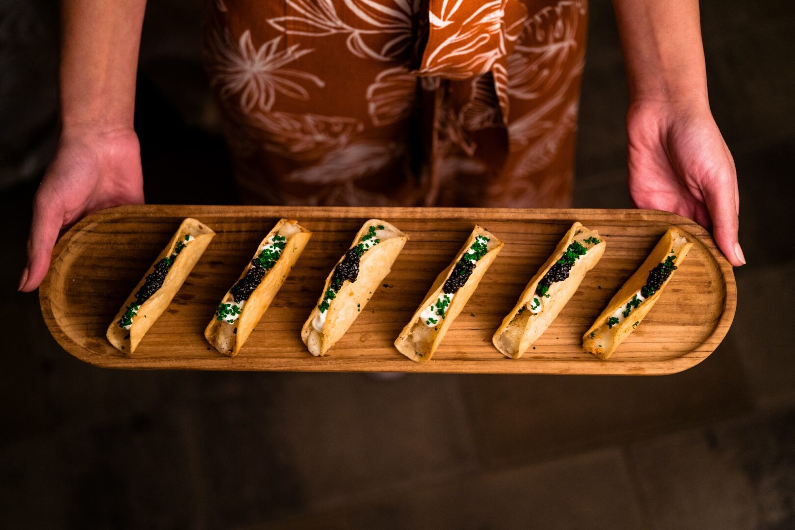 A person holds a wooden tray with six neatly arranged appetizers topped with black and green garnishes. The person wears a brown, floral-patterned outfit.