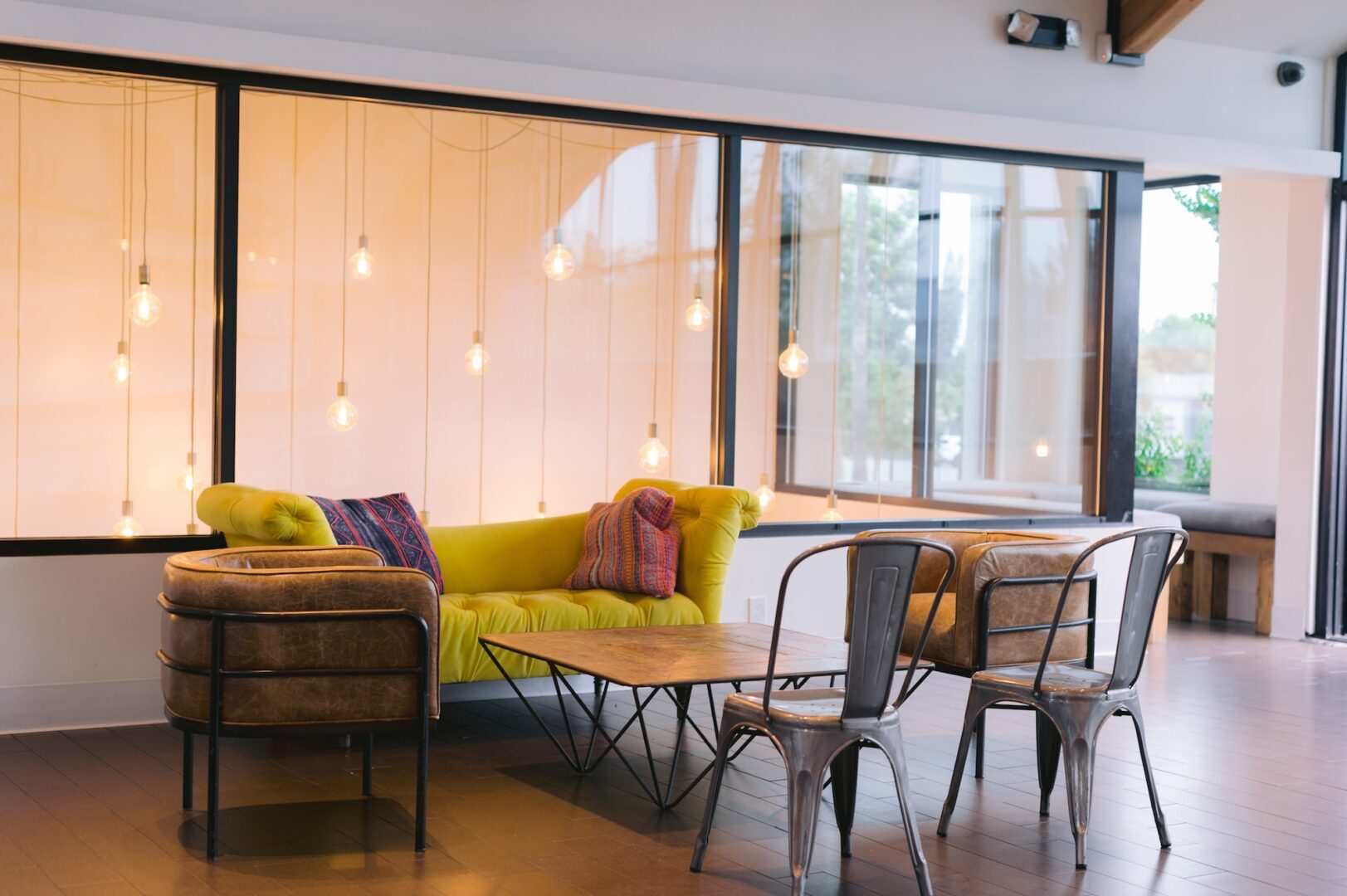 A cozy lounge area features a bright yellow couch with pillows, a wooden coffee table, two metal chairs, and hanging lightbulbs in front of large windows.