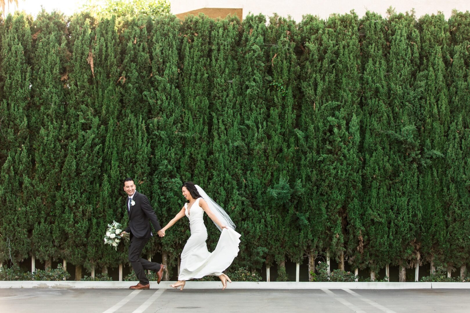 A bride and groom joyfully run barefoot together in a parking lot, holding hands. The bride lifts her dress, and the groom holds a bouquet. Tall, dense green trees fill the background.