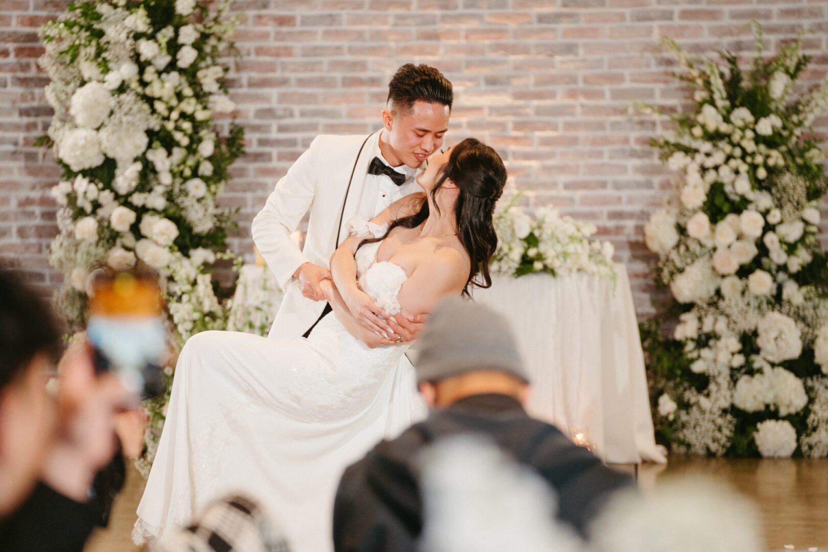 A groom in a white tuxedo dips and kisses his bride during their wedding, surrounded by white flowers and greenery, with guests taking photos in the foreground.