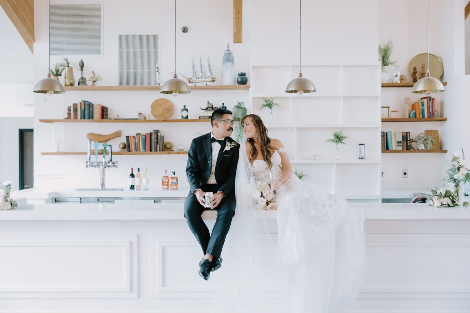 A bride and groom sit on a modern white counter, smiling at each other in a bright room with shelves of books and decor behind them.
