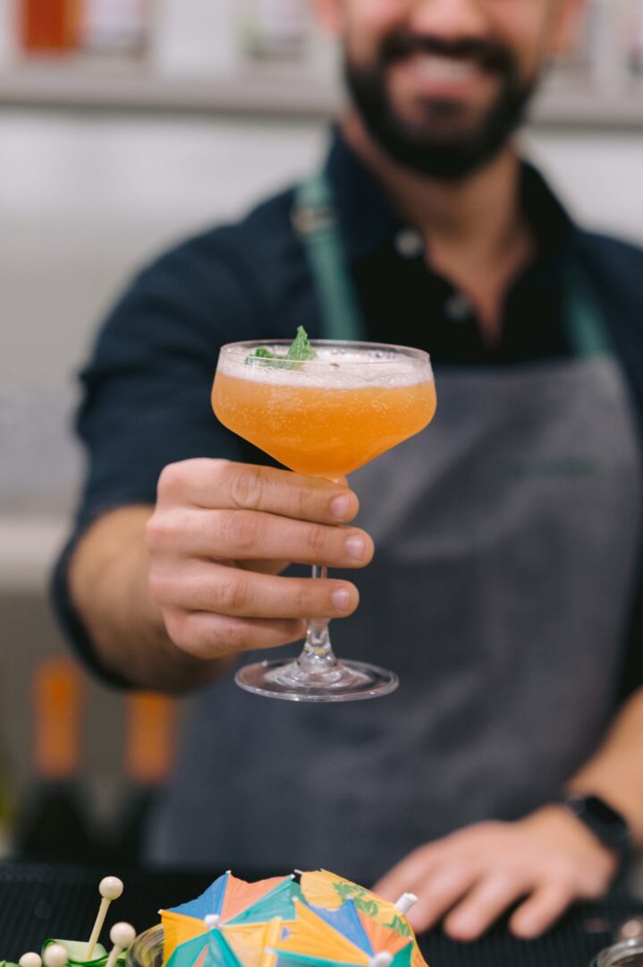 A bartender in a gray apron smiles while holding out an orange cocktail in a coupe glass, garnished with a mint leaf. A colorful paper umbrella decoration is visible in the foreground.