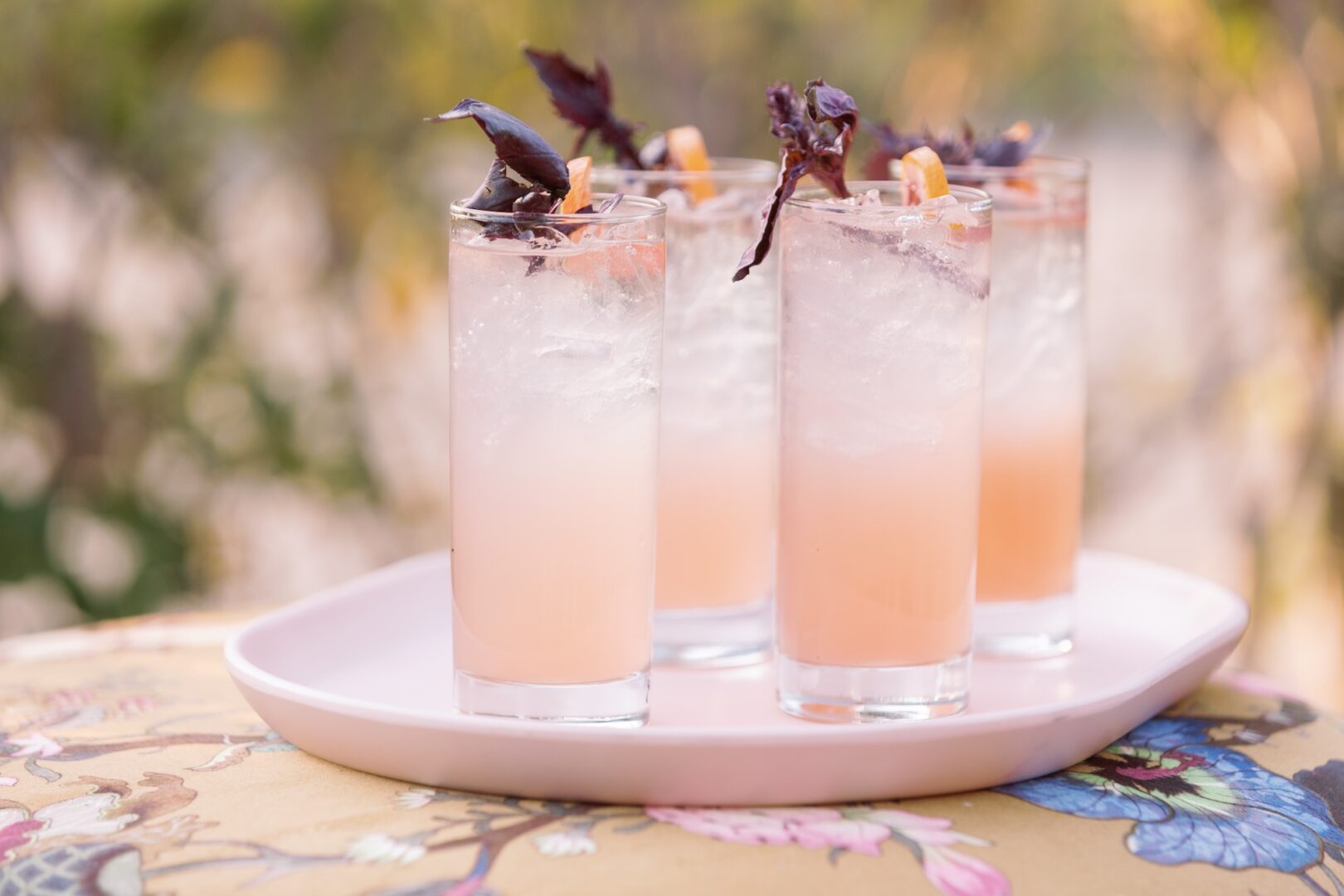 Four tall glasses of a light pink drink with ice, garnished with dark purple basil and orange peel, are arranged on a white tray atop a floral-patterned tablecloth.
