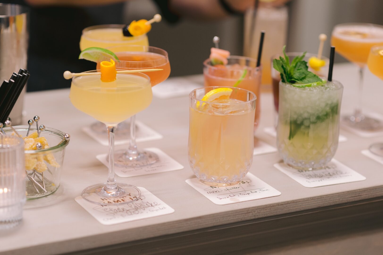 A row of colorful cocktails in various glasses, each garnished with fruit or herbs, sits on coasters labeled with handwritten drink names on a bar counter.