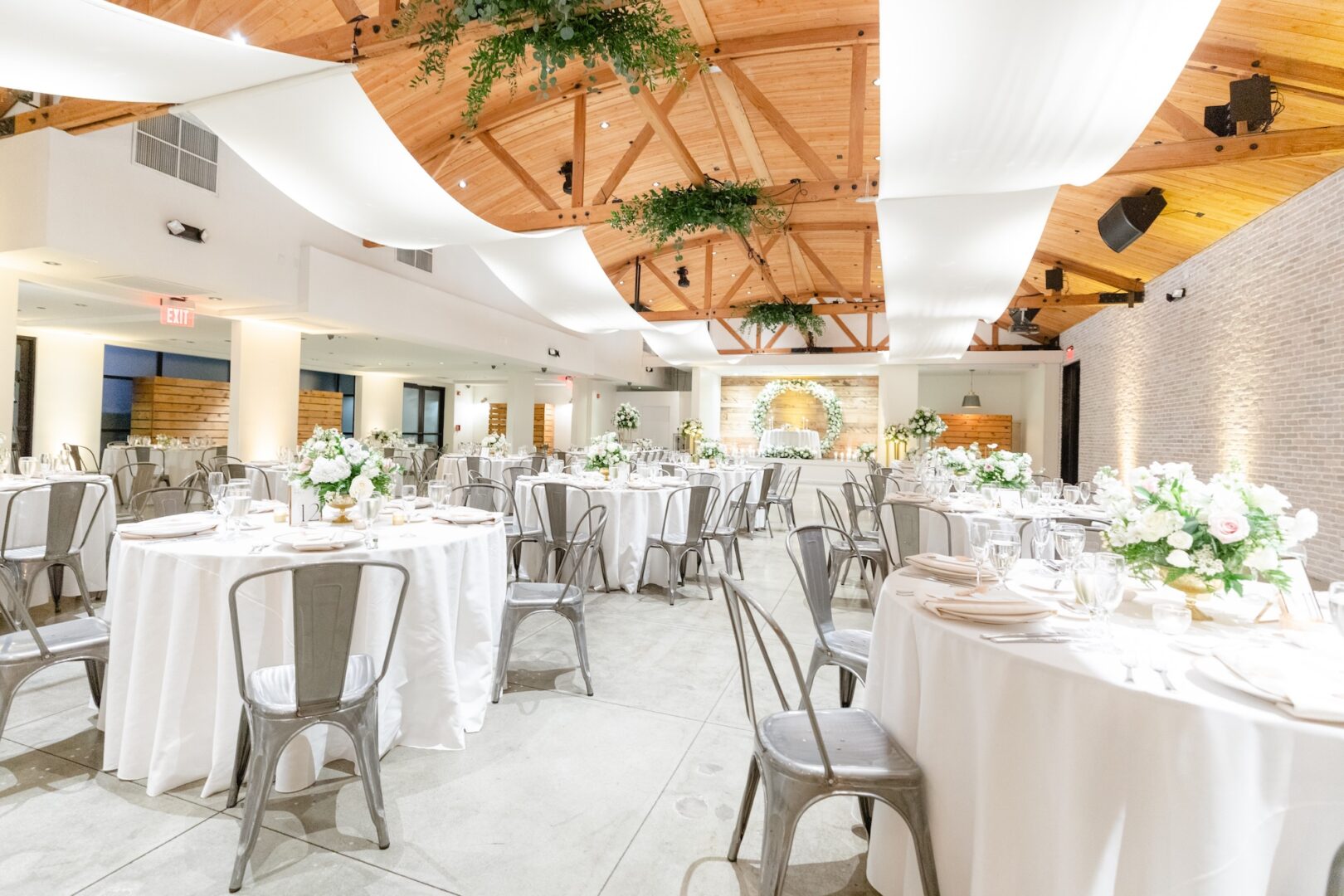 A bright, elegant event hall with round tables set for a formal occasion. White tablecloths, floral centerpieces, and metal chairs fill the space under wooden beams and draped white fabric.
