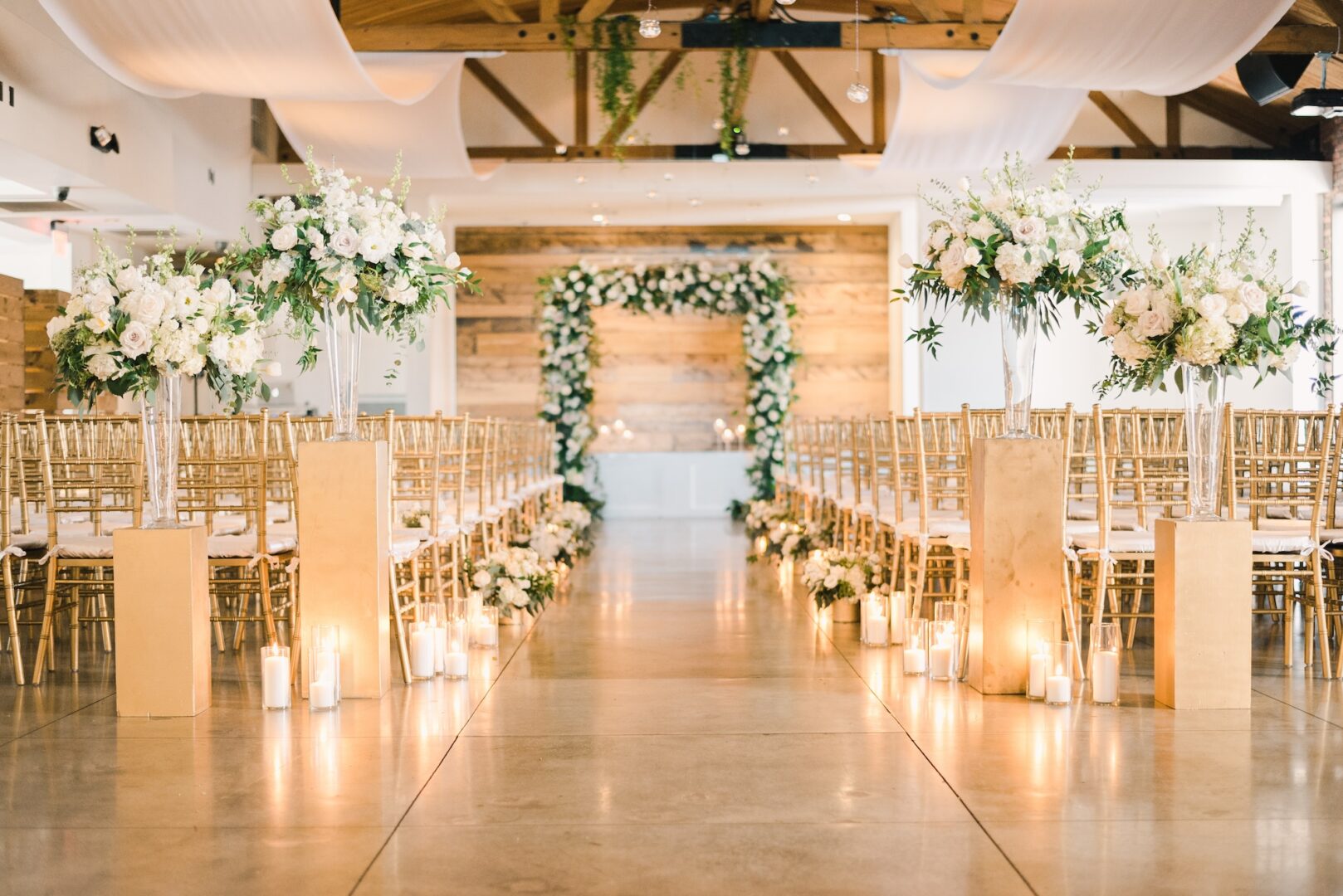 Elegant indoor wedding ceremony setup with gold chairs, white floral arrangements, candles lining the aisle, and a wooden arch adorned with white flowers and greenery at the front. Warm lighting creates a romantic atmosphere.