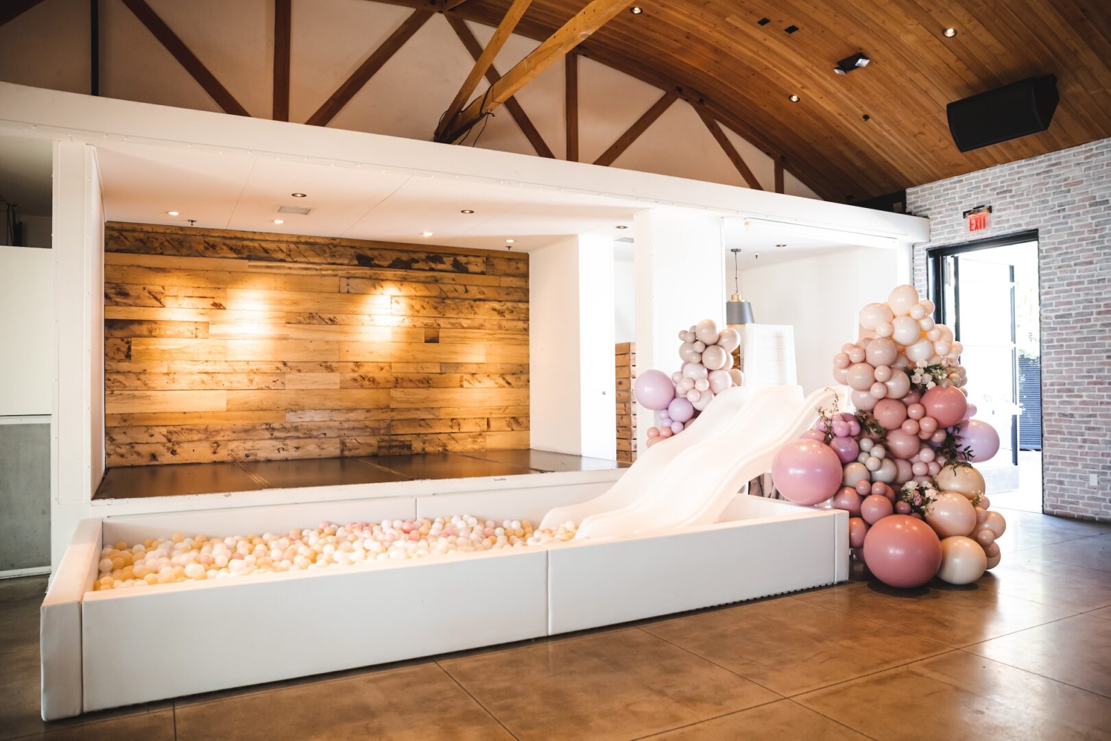 A modern indoor play area with a white slide leading into a ball pit, surrounded by pastel-colored balloon decorations. The space has wooden accents and exposed beams.