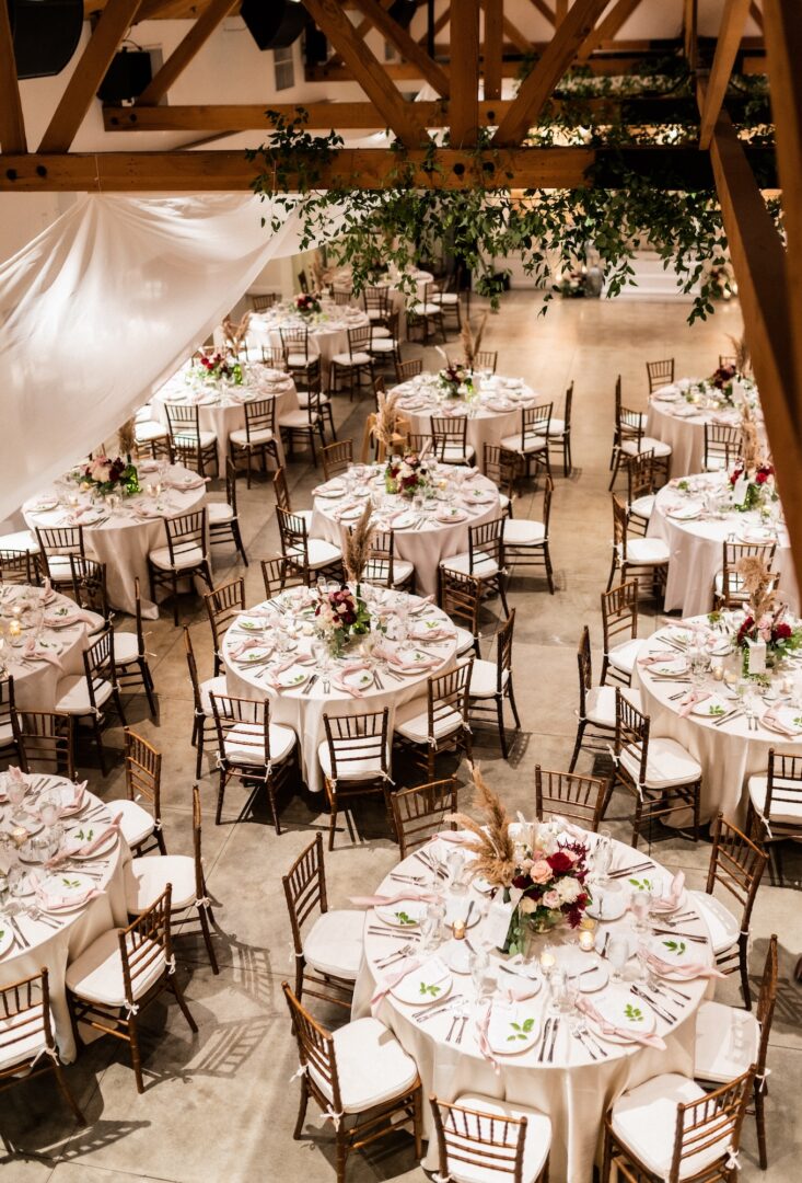 A wedding reception hall with round tables set for guests, decorated with flower centerpieces and white linens, wooden chairs, and greenery hanging from wooden ceiling beams.