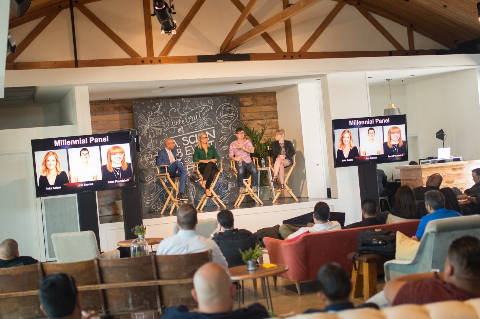 A panel of four people speaks on stage in front of an audience. Two screens display Millennial Panel with headshots and names. The setting is a modern, open room with wood beams and a chalkboard backdrop.