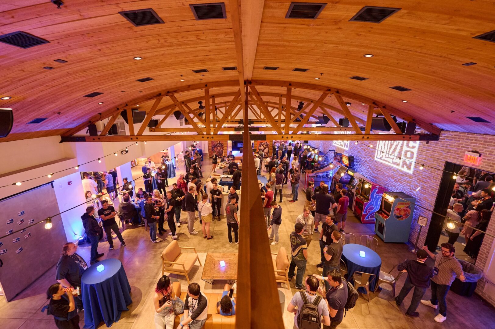A large group of people mingle at an indoor event with a wooden arched ceiling. There are arcade machines, tables with blue tablecloths, and soft lighting throughout the space.