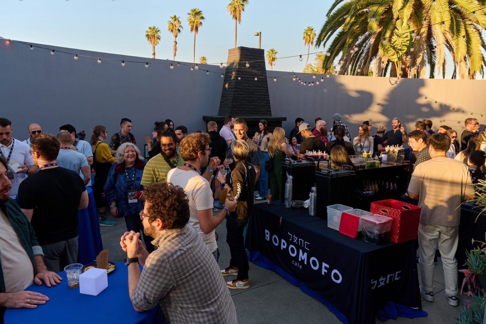 A diverse crowd enjoys an outdoor social event with drinks and food at sunset; people mingle around tables under string lights, palm trees, and clear skies. A Bopomofo Cafe booth is visible in the foreground.