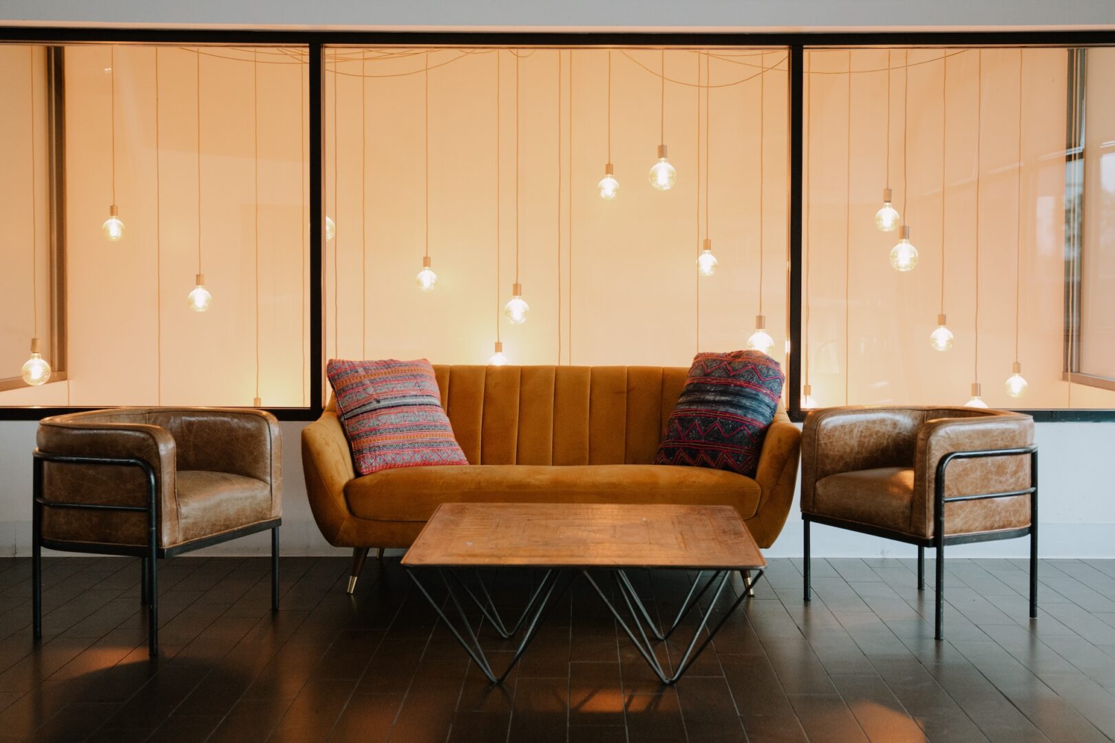 A modern living room features a mustard sofa with two colorful pillows, two brown armchairs, a wooden coffee table, and hanging lightbulbs in front of large windows with an orange tint.