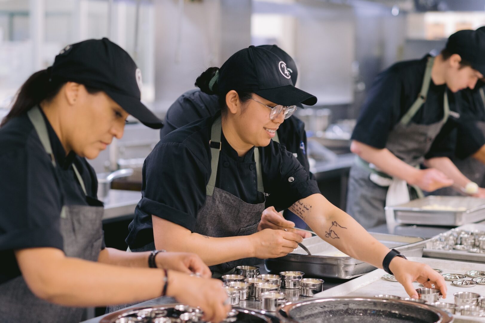 Three chefs in black uniforms and hats prepare food at a stainless steel counter in a commercial kitchen, handling small metal containers and smiling while working together.