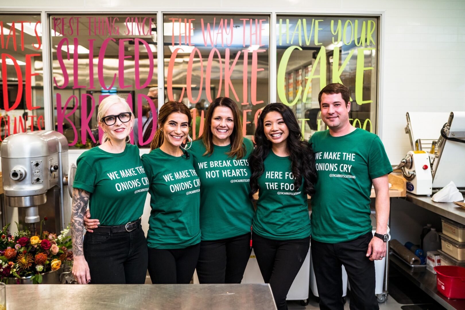 Five people in matching green shirts with baking-related slogans stand smiling in a bright kitchen with colorful writing on the windows behind them. Mixing equipment and flowers are visible on the counter nearby.