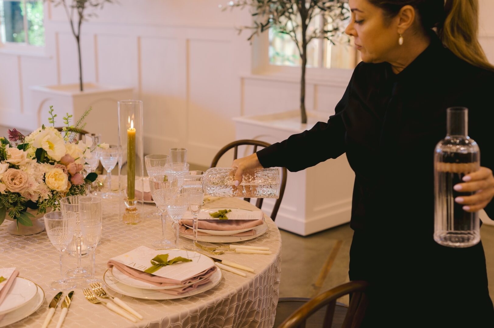 A woman pours water into a glass at an elegantly set round table with floral centerpiece, candles, and neatly arranged plates, glasses, and cutlery.