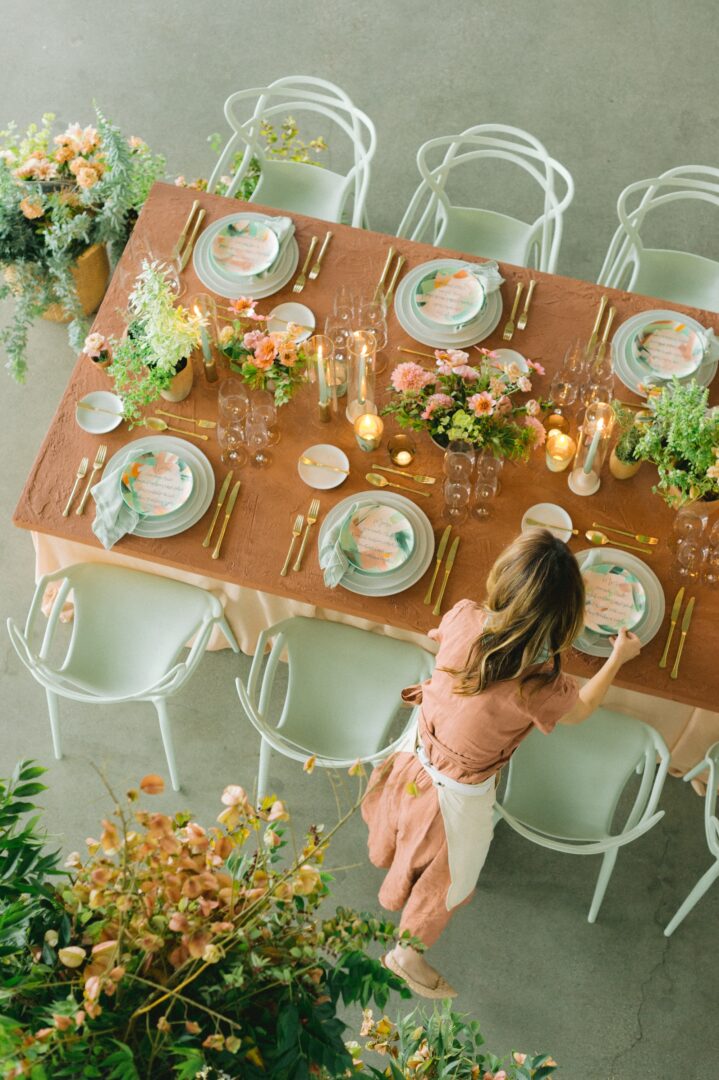 A person arranges a beautifully set dining table with floral centerpieces, candles, and elegant place settings, viewed from above. Light green chairs surround the table.