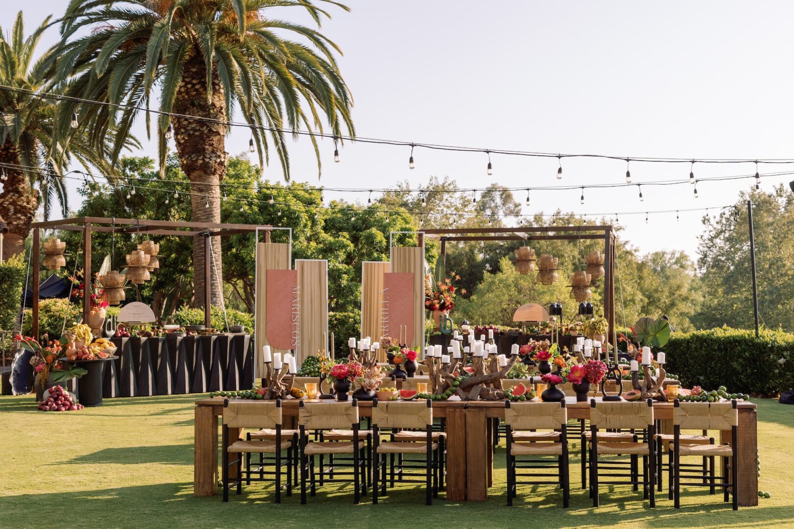 Outdoor dining setup on grass with wooden tables, chairs, colorful flowers, hanging lanterns, string lights, palm trees, and lush greenery in the background under a clear sky.