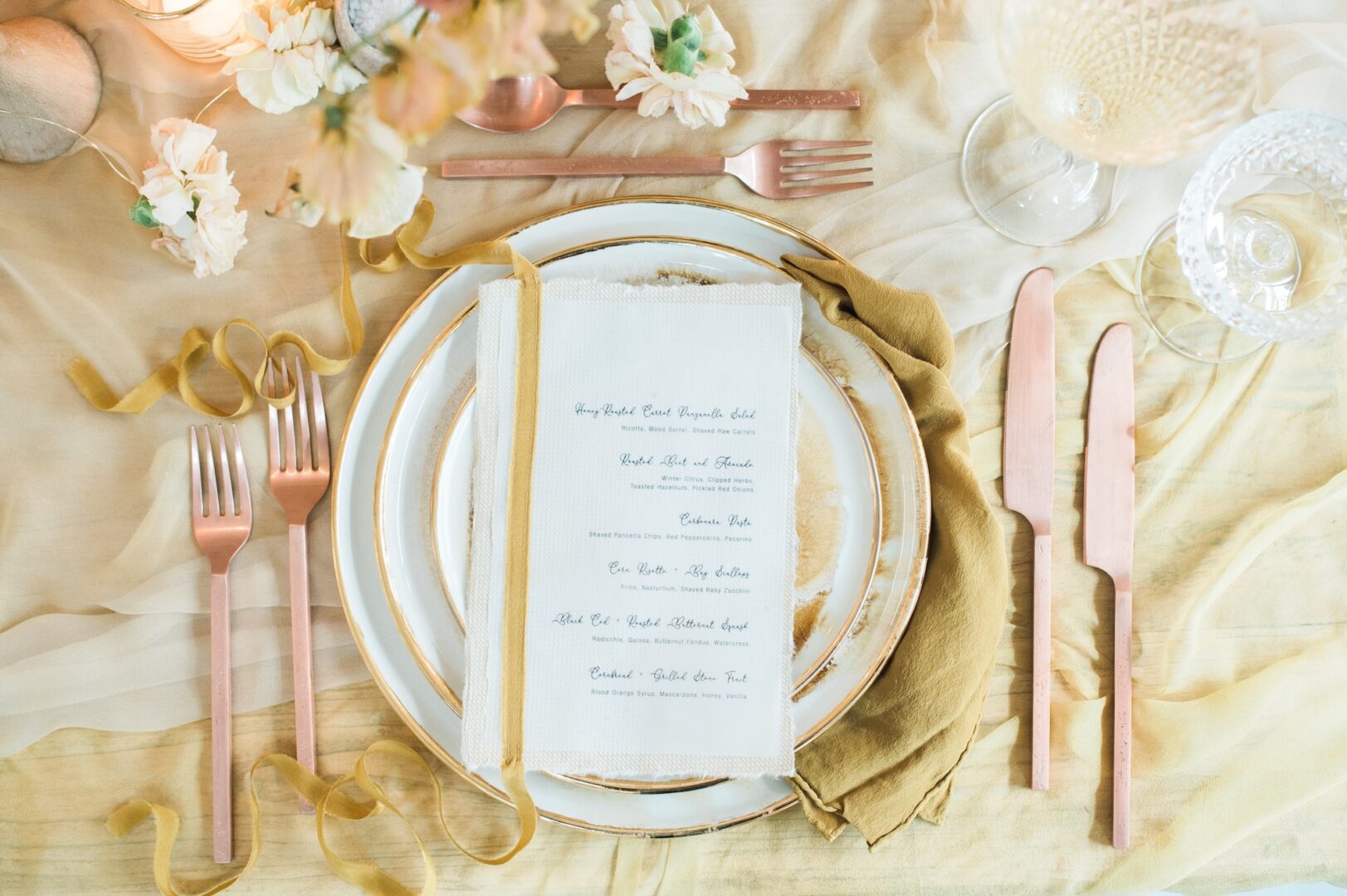 Elegant table setting with gold and blush pink cutlery, layered plates, a gold napkin, and a printed menu on white fabric. Flowers and glassware complete the sophisticated, romantic atmosphere.