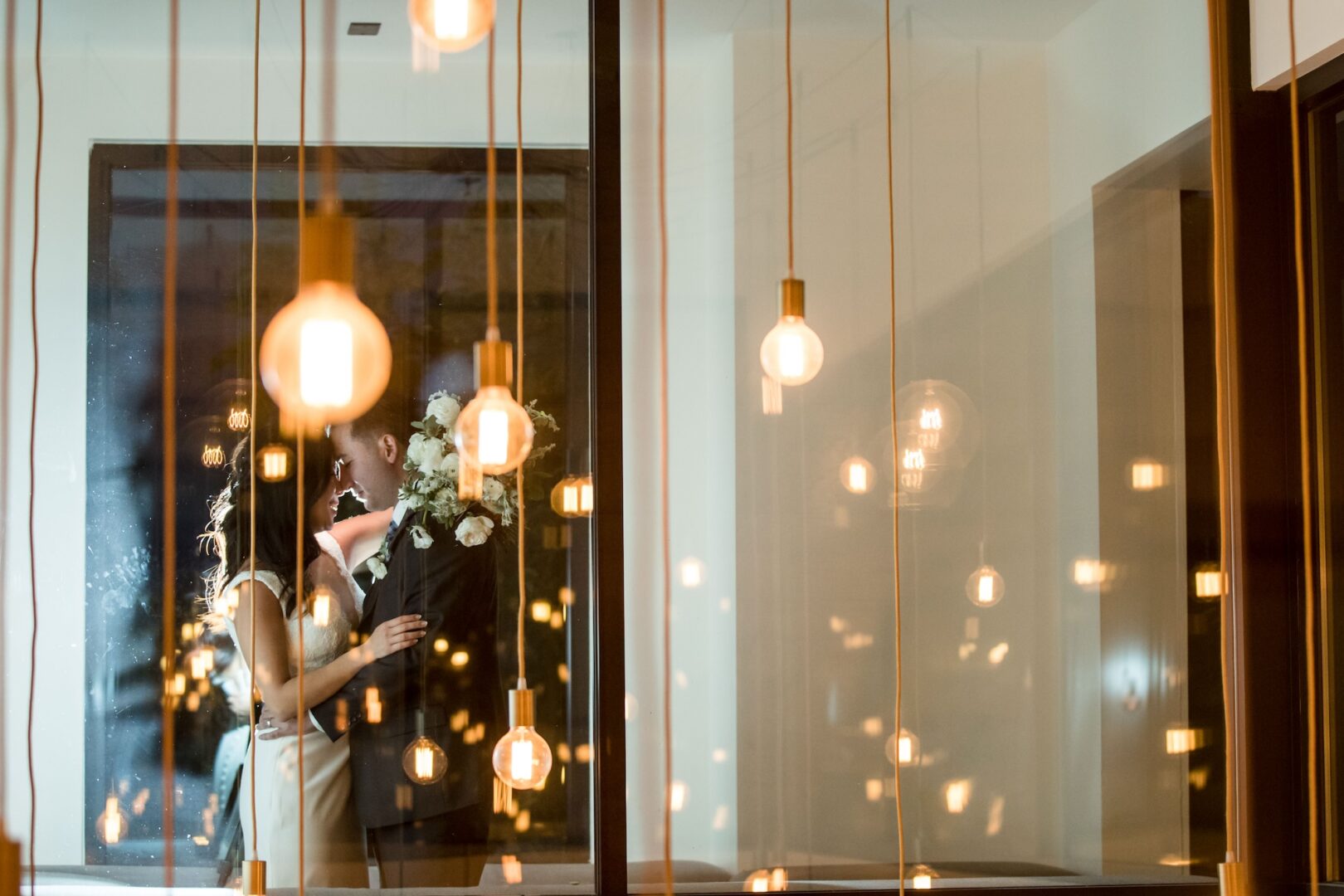 A bride and groom embrace and kiss behind a glass panel decorated with hanging lightbulbs, creating a romantic and dreamy atmosphere.