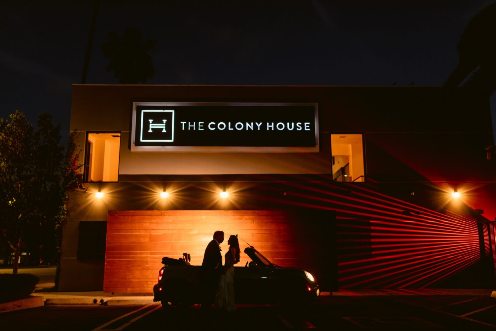 A couple stands in silhouette by a convertible car at night, in front of The Colony House building, illuminated by warm exterior lights.