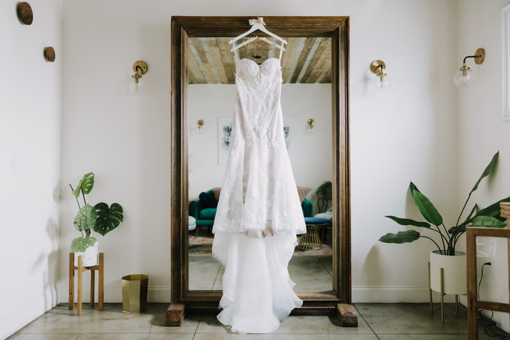 A white lace wedding dress hangs on a wooden-framed mirror in a bright room with plants, sconces, and a tiled floor. A green couch and framed art are reflected in the mirror behind the dress.