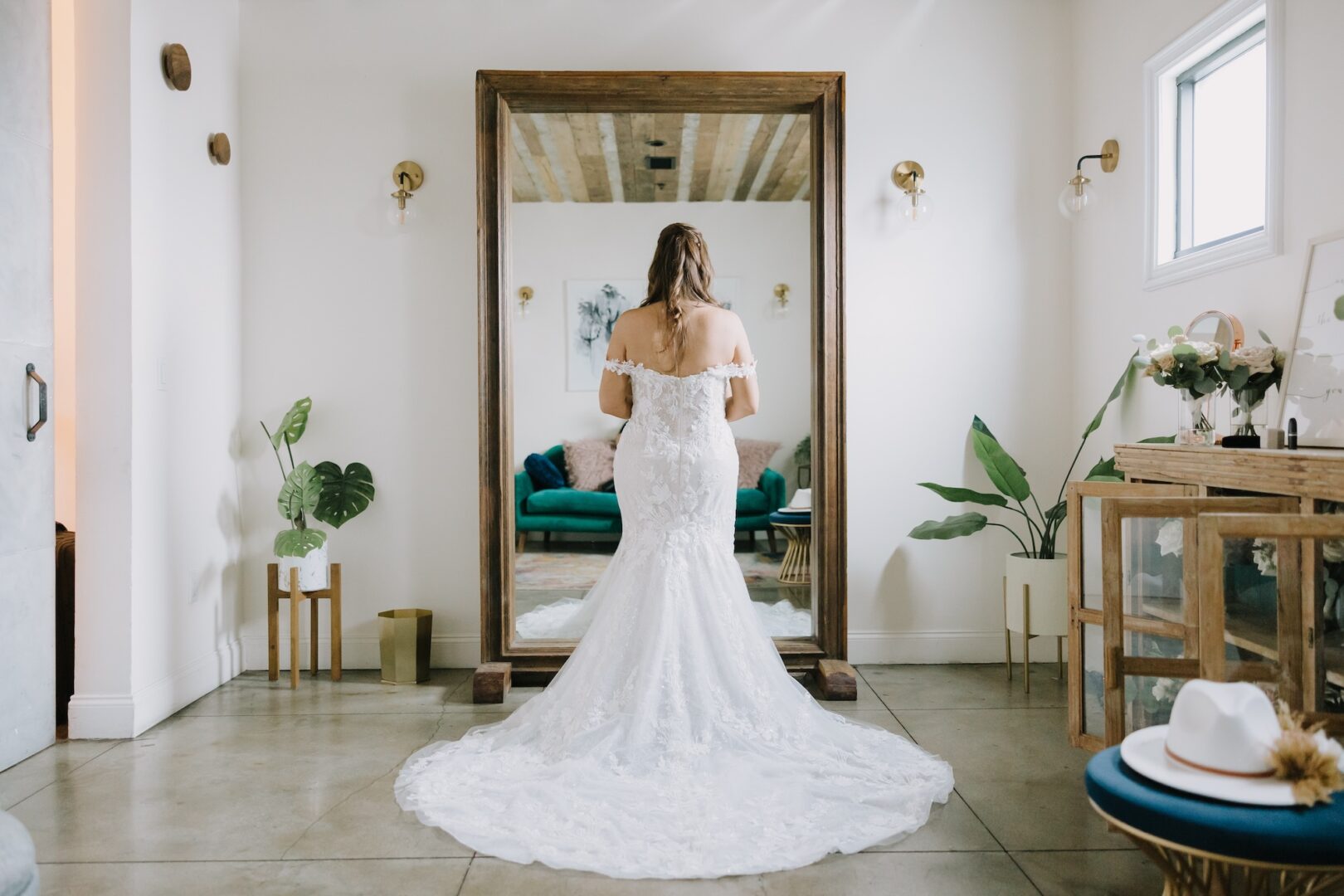 A bride in a white lace gown stands facing a tall mirror in a stylish, modern room with plants, gold accents, and natural light.