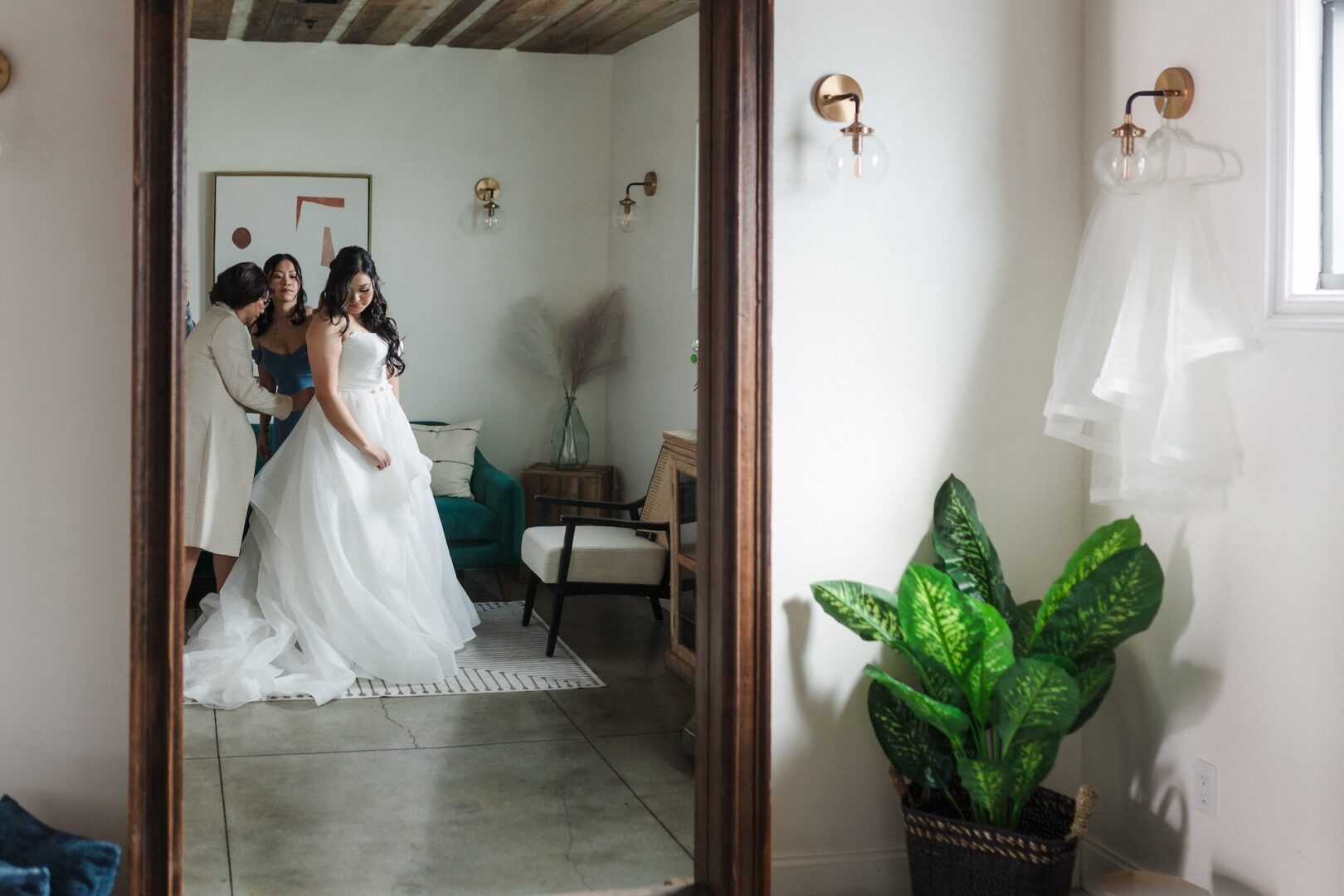 A bride in a white gown stands with two women helping her, reflected in a large mirror inside a modern, softly lit room with plants and simple decor.