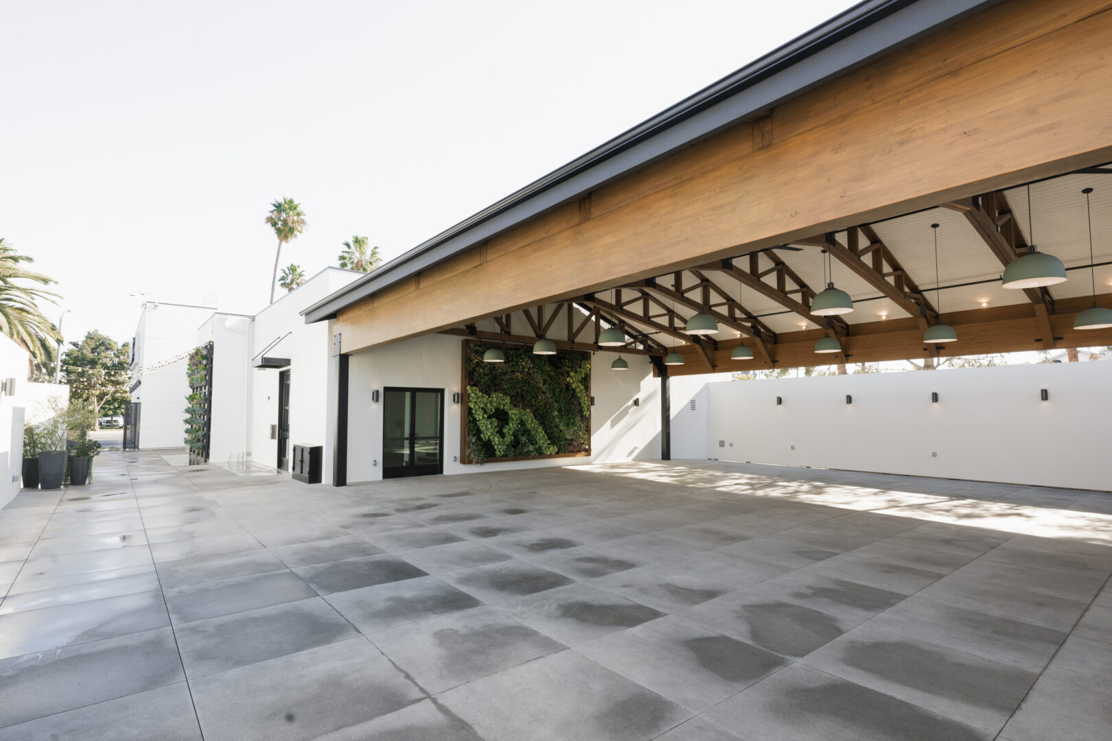 Modern open-air pavilion with exposed wooden beams, hanging lights, and a vertical garden on one wall. The spacious concrete floor is partially wet, and palm trees are visible in the background outside the structure.