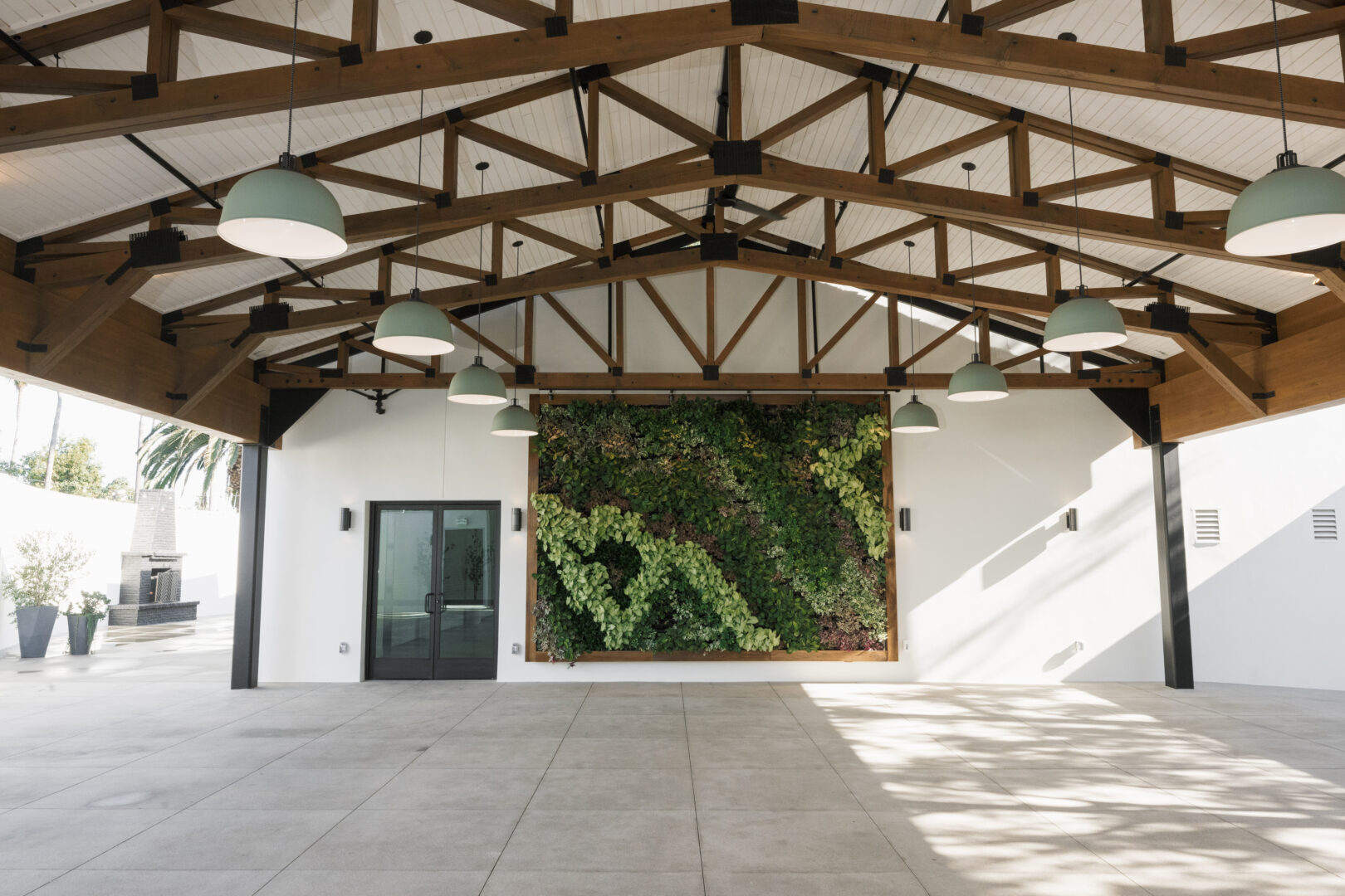 Open pavilion with exposed wooden beams, large hanging lights, a living green wall in the center, a black-framed glass door on the left, and a white concrete floor.
