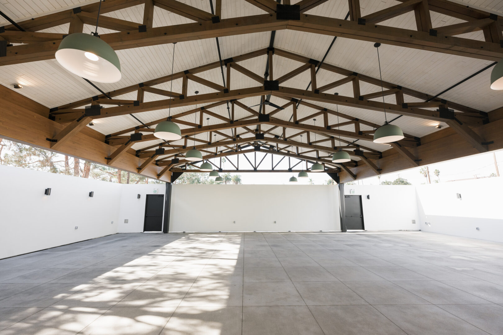 Spacious, empty indoor area with high wooden beams, large hanging lights, white walls, tiled floor, and natural light casting shadows through windows.