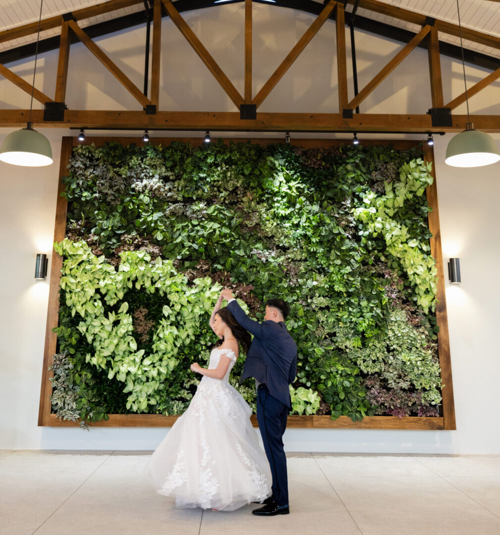A bride and groom dance together indoors in front of a large green living plant wall with wooden beams and overhead lights.
