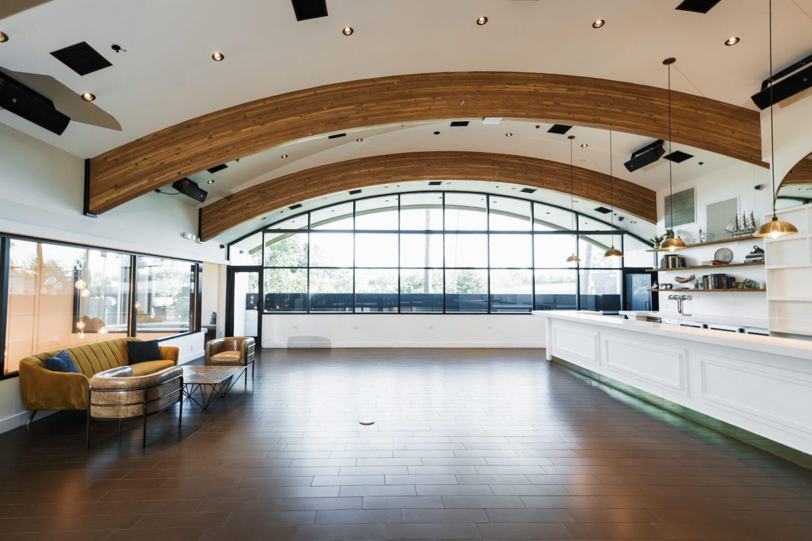Spacious modern room with arched wooden beams, large windows, a small seating area with two armchairs and a couch, and a white counter with gold pendant lights.
