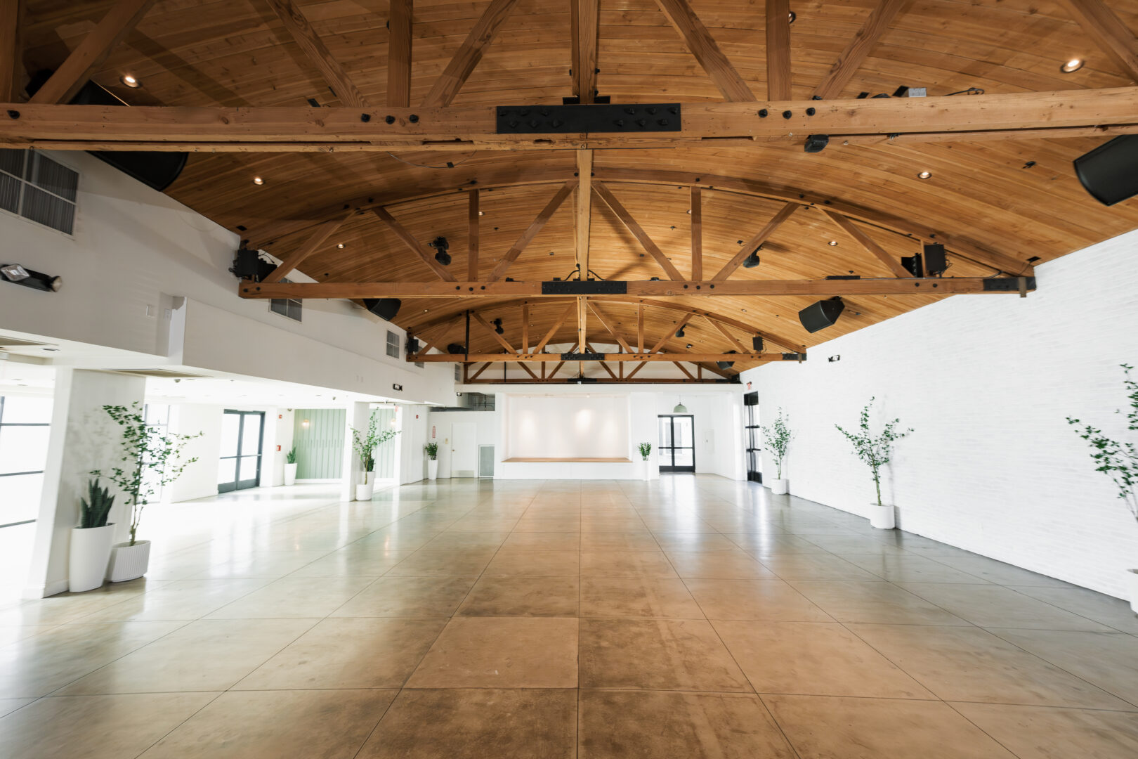 Spacious, empty event hall with a wooden vaulted ceiling, white walls, large windows, potted plants, and a small stage at the far end. Natural light fills the room.