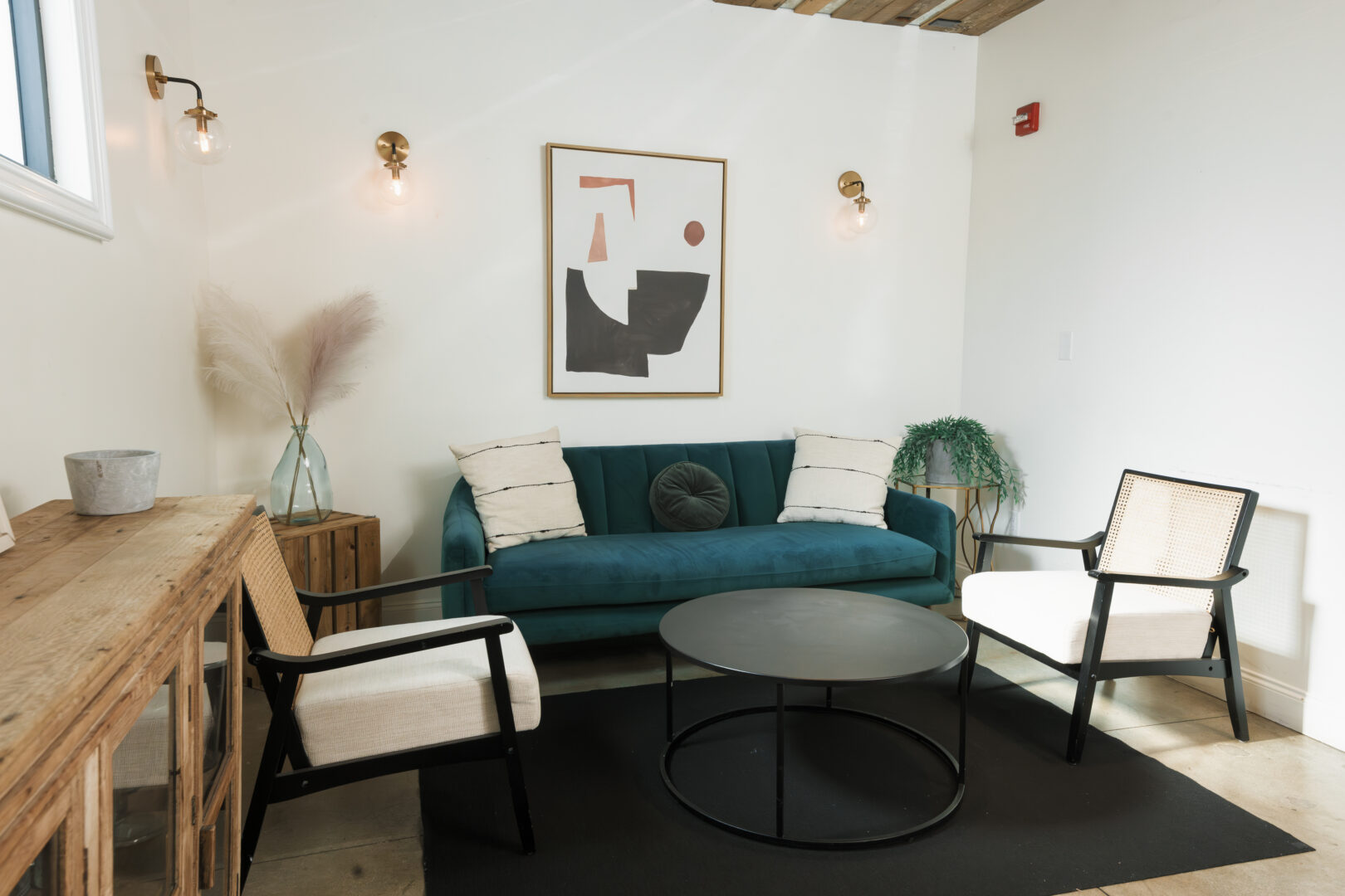A cozy living room features a teal sofa, two cream armchairs, a round black coffee table, abstract wall art, potted plants, and soft lighting against white walls and a wooden ceiling accent.