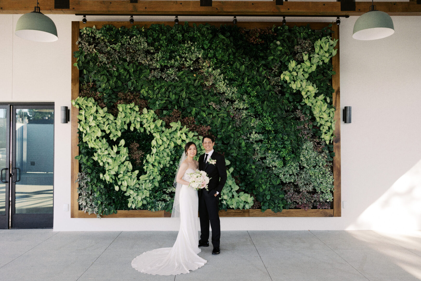 A bride and groom stand together smiling in front of a large green plant wall, with the bride holding a white bouquet and both dressed in formal wedding attire.