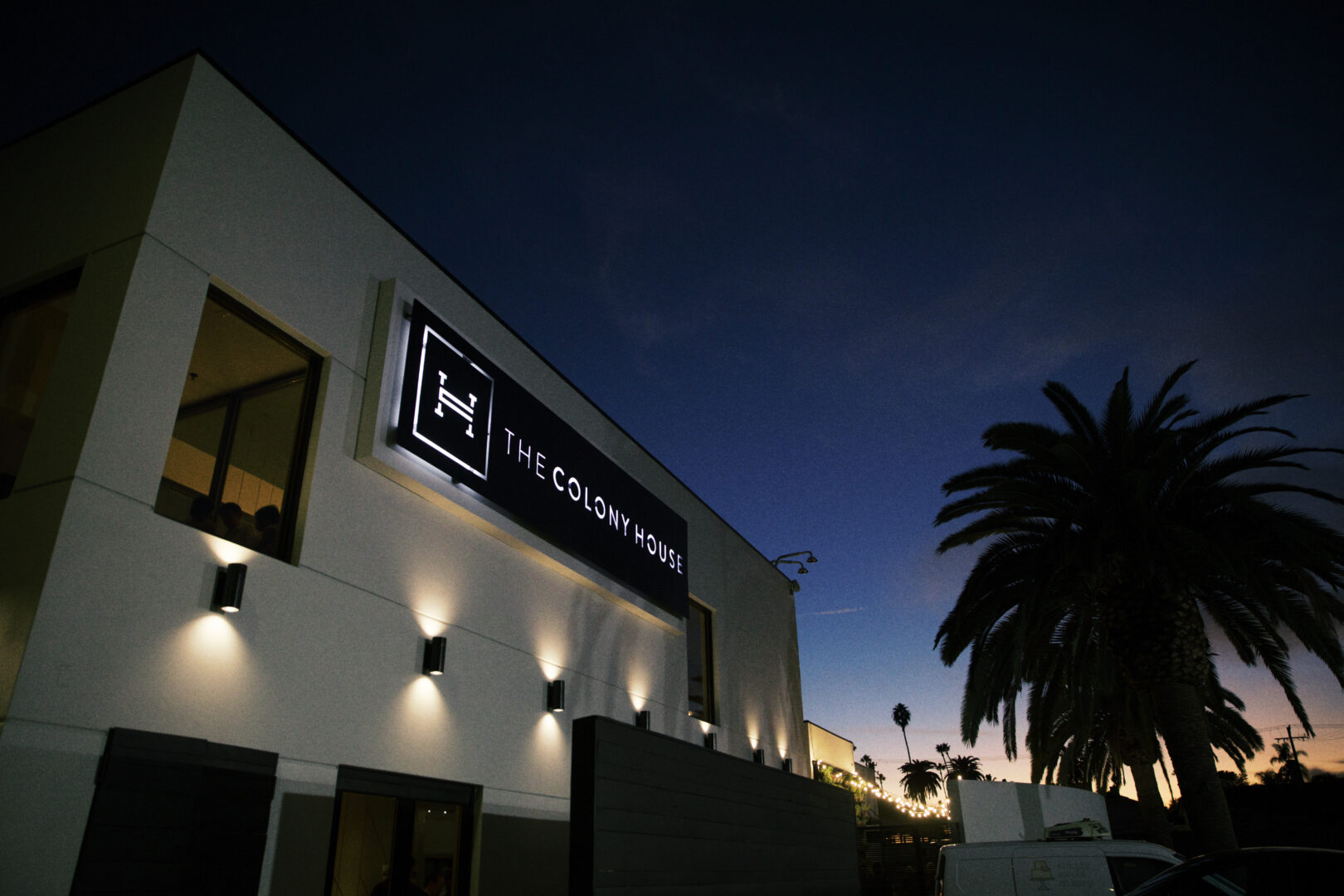 A modern building with a lit sign reading The Colony House at dusk, palm trees and a darkening sky in the background.