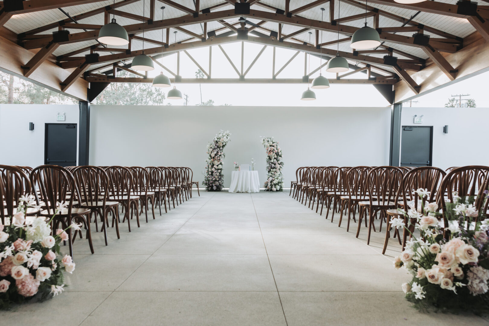 Rows of brown chairs face a white altar with floral arches and arrangements in a bright, modern indoor wedding venue with high ceilings and exposed beams.