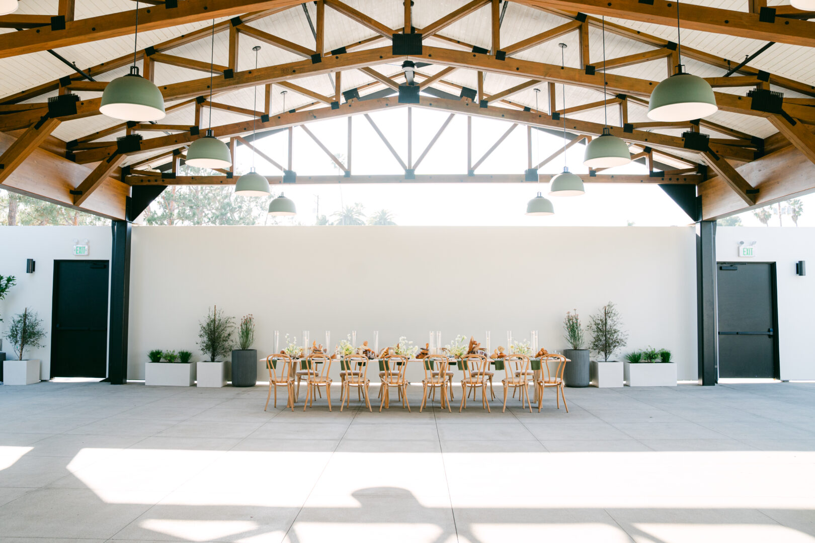 A long table set for a meal with wooden chairs sits under a modern pavilion with exposed beams, large hanging lights, and an open, airy space.