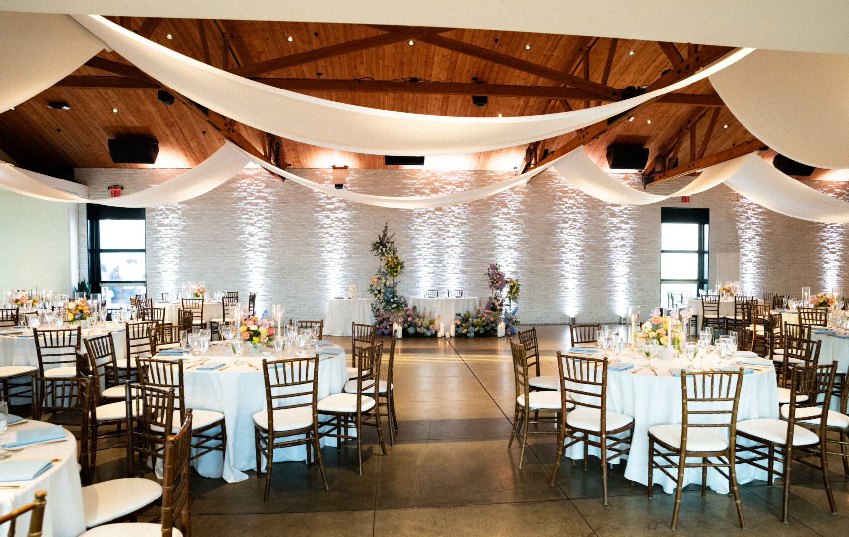 Elegant wedding reception hall with round tables covered in white linens, wooden chairs, floral centerpieces, and draped fabric across a wooden-beamed ceiling. A decorated backdrop is set up at the front.