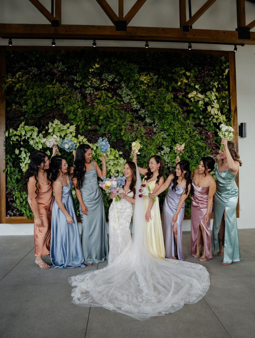 A bride in a white dress stands with seven bridesmaids in colorful satin dresses, all smiling and laughing in front of a lush green wall with flowers.