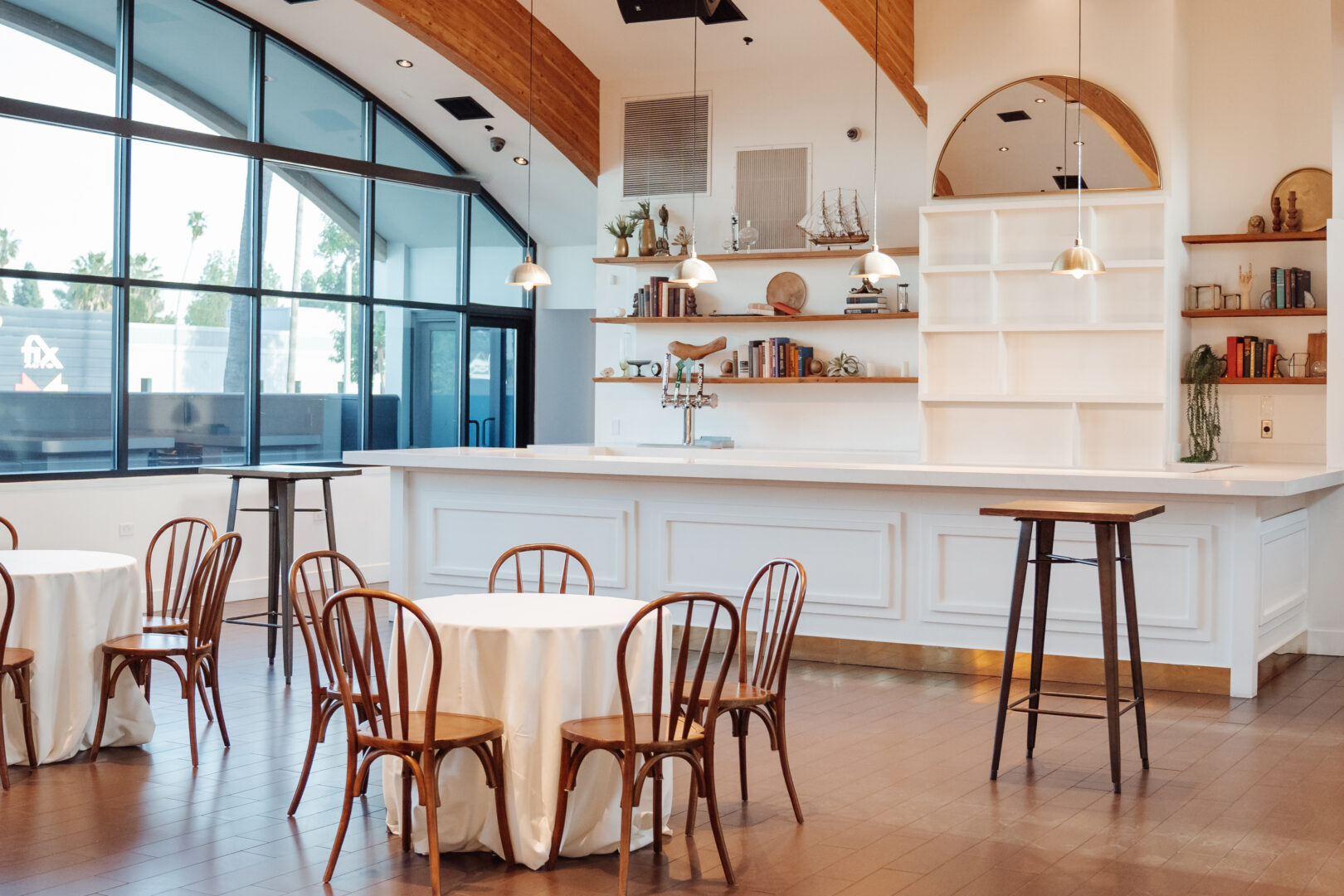 Bright, modern café with round tables covered in white linens, wooden chairs, a large white counter, open shelves with décor, pendant lighting, and large arched windows letting in natural light.