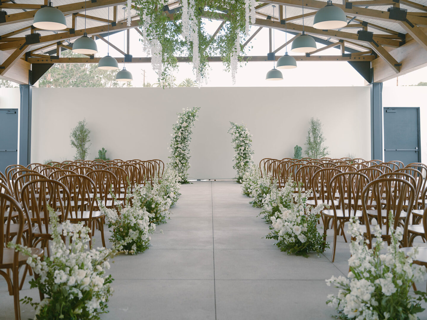 A modern indoor wedding venue with wooden chairs arranged in rows and white flowers lining the aisle. Greenery hangs from the ceiling, and floral arches stand at the front.